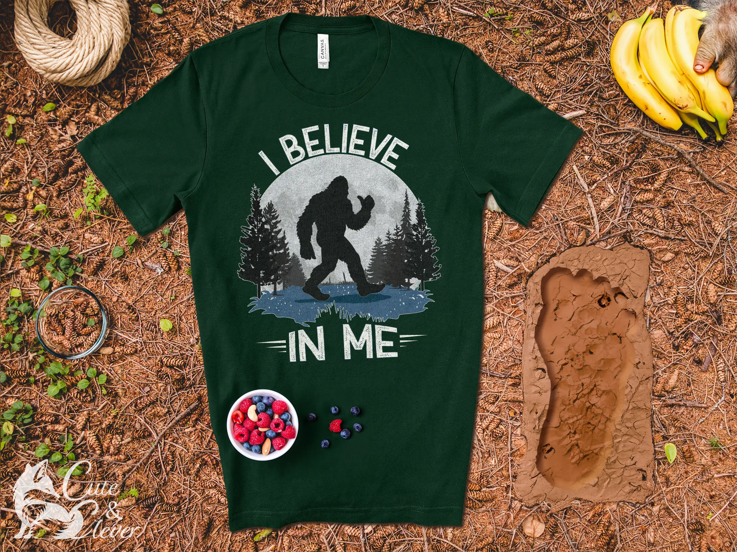 A Bigfoot Believes T-Shirt lays on a bed of pine needles, a footprint, bananas and a bowl of nuts and berries nearby.