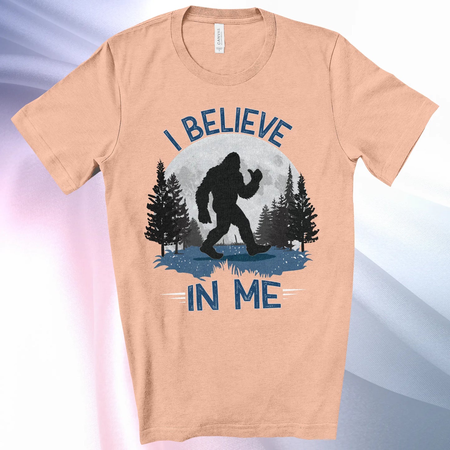 Heather Peach Bigfoot Believes T-Shirt with Bigfoot's silhouette in a moonlit forest and I Believe at the top and In Me below