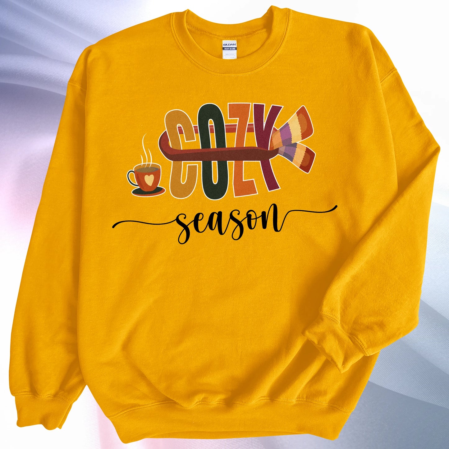 Gold colored Cozy Season sweatshirt, on colorful gradient silk background. Left sleeve folded over the garment at the elbow