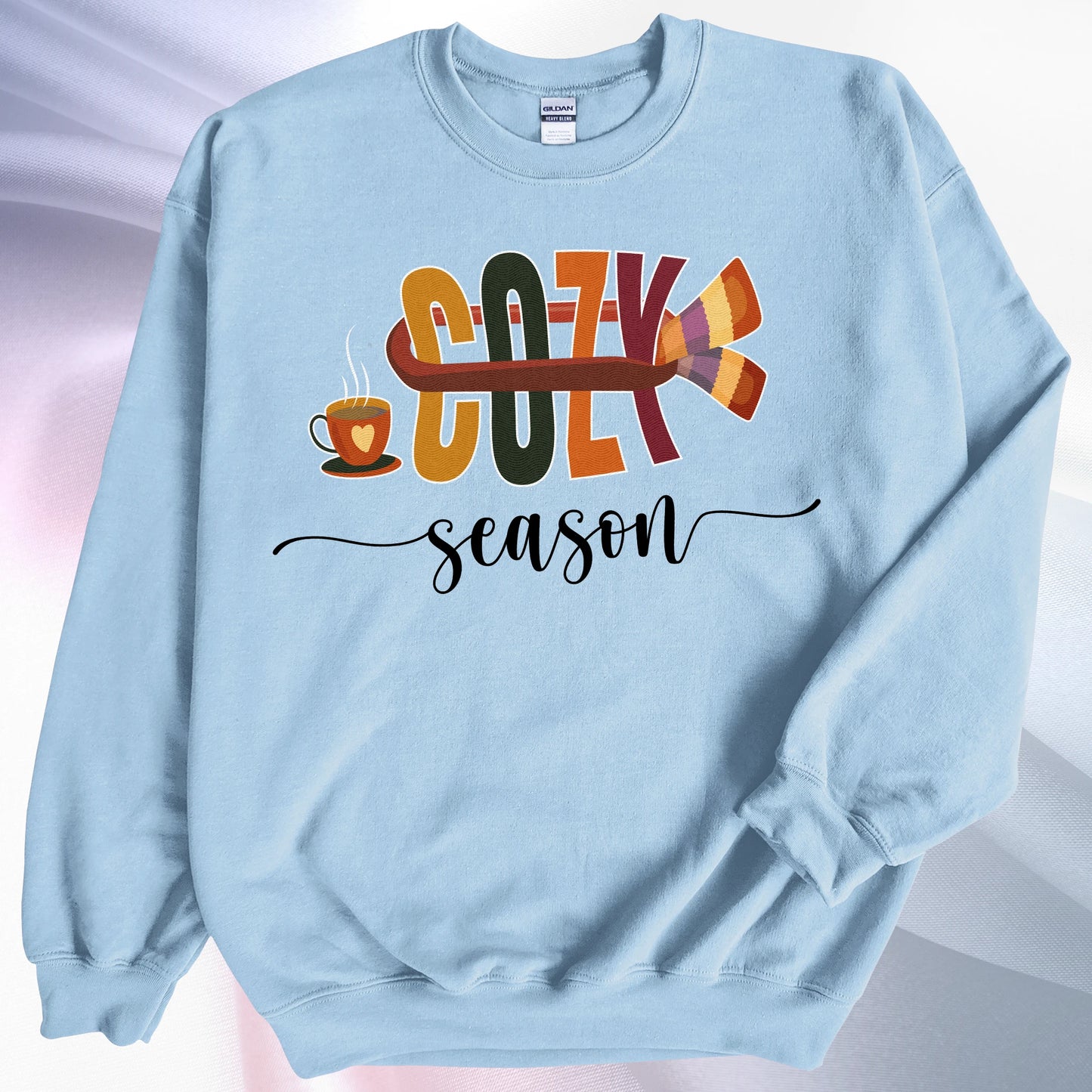 Light blue Cozy Season sweatshirt, on colorful gradient silk background. Left sleeve is folded over the garment at the elbow