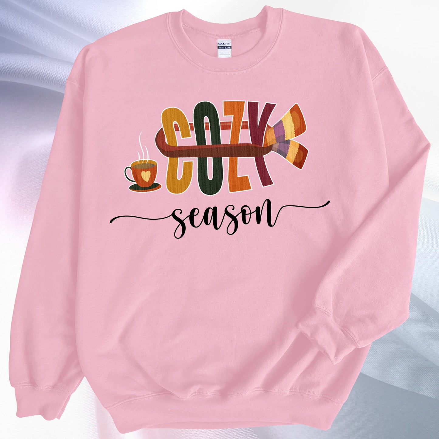 Light pink Cozy Season sweatshirt, on colorful gradient silk background. Left sleeve is folded over the garment at the elbow