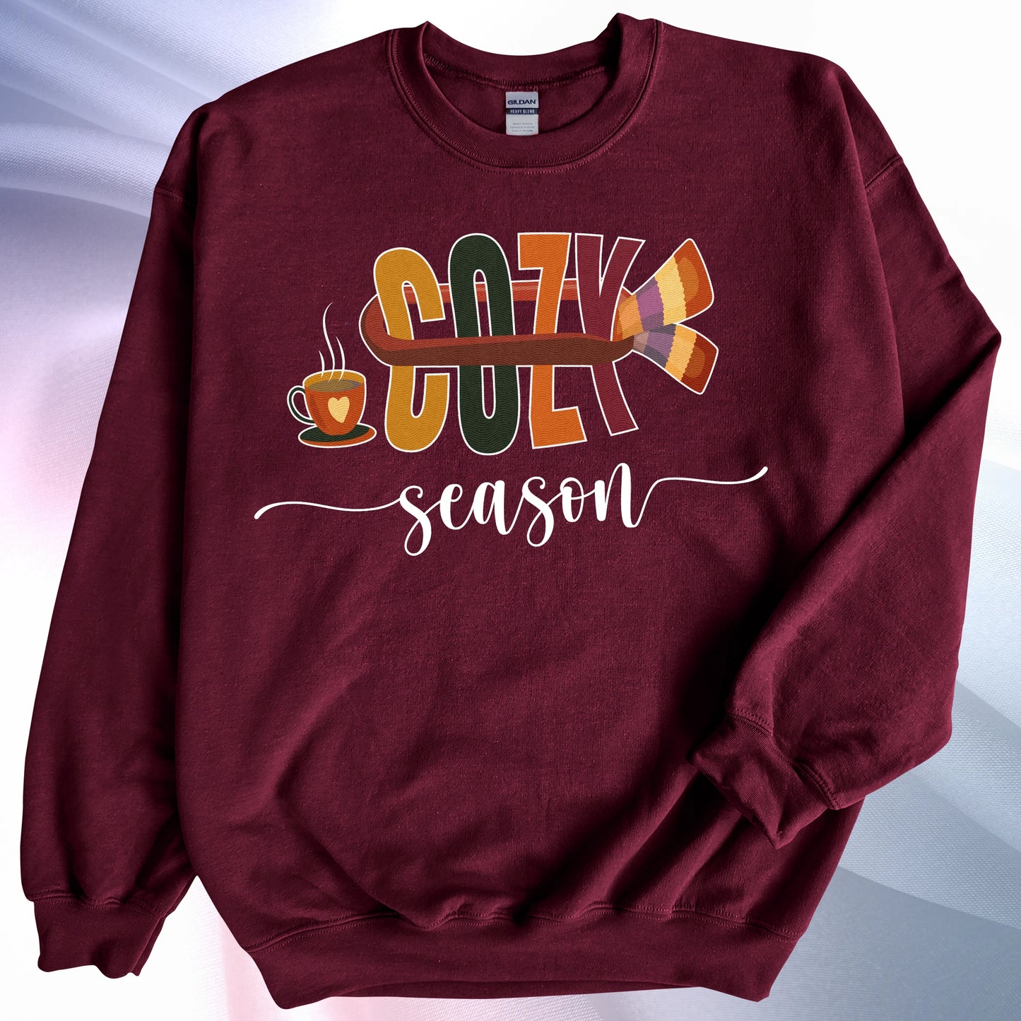 Maroon colored Cozy Season sweatshirt, on colorful gradient silk background. Left sleeve folded over the garment at the elbow