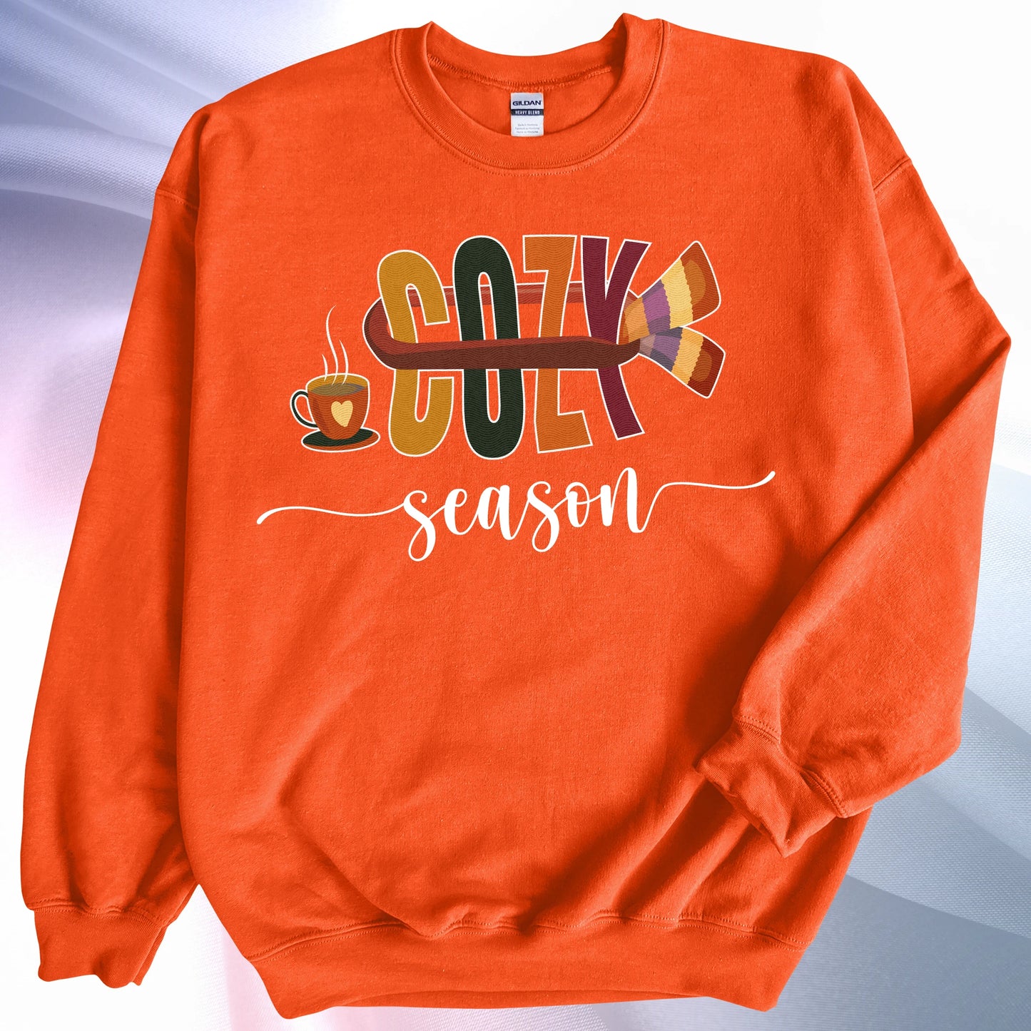 Orange Cozy Season sweatshirt, placed on colorful gradient silk background. Left sleeve folded over the garment at the elbow