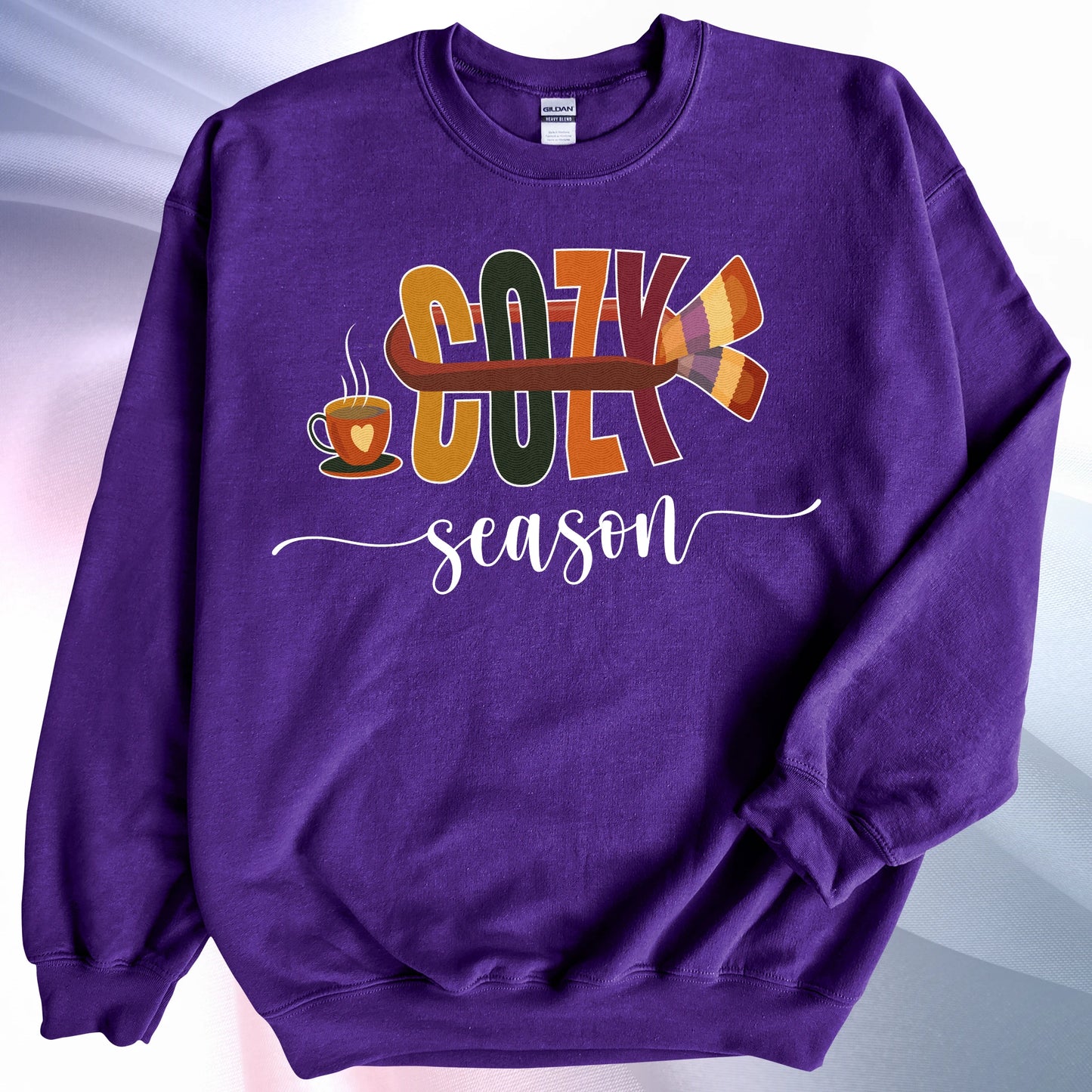 Purple Cozy Season sweatshirt, laying on colorful gradient silk background. Left sleeve folded over the garment at the elbow