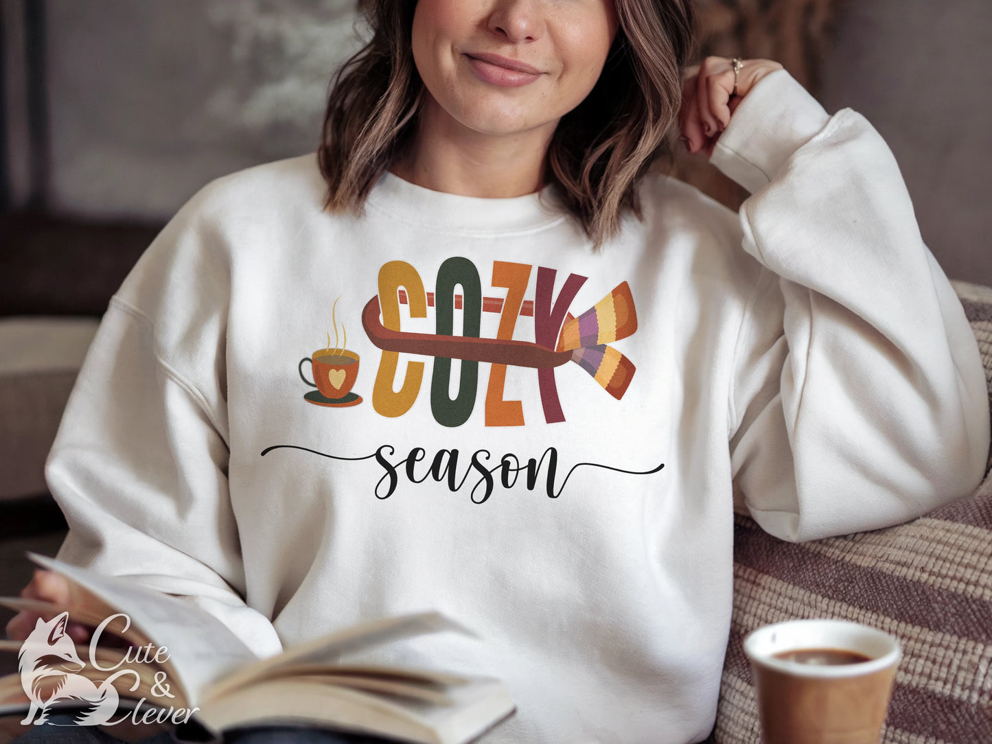 Relaxed, smiling female model wearing white Cozy Season sweatshirt sits on a comfy couch while reading and drinking coffee
