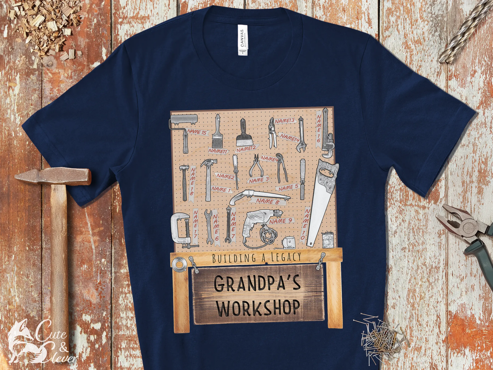 A Navy colored Grandpa's Workshop T-Shirt lays on a rustic wooden table with pliers, drill bits, a hammer, nails and wood chips.