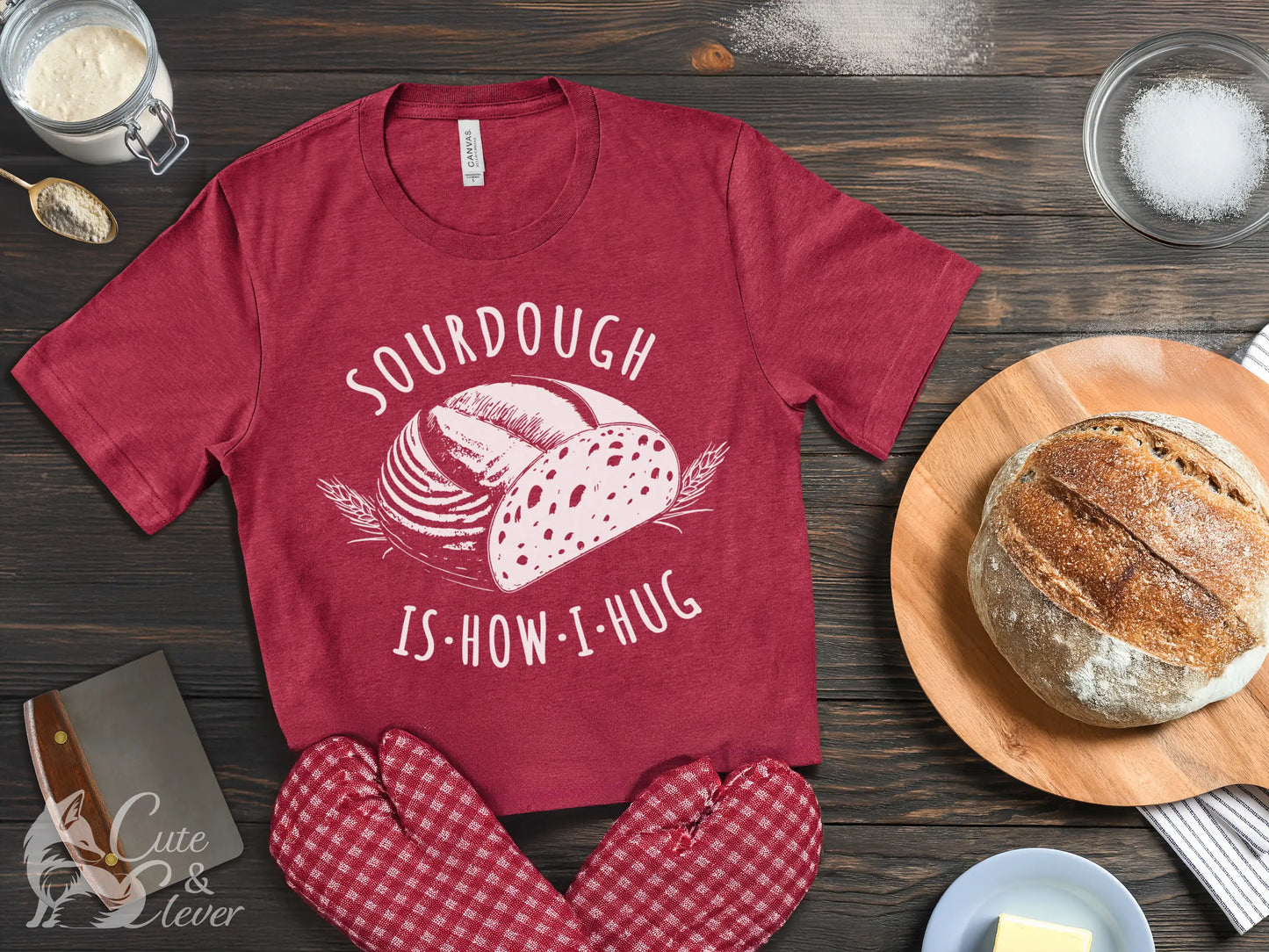 A Heather Raspberry colored Sourdough Hug T-Shirt lays on a wooden table with starter, salt, sourdough, dough scraper, mittens and butter.