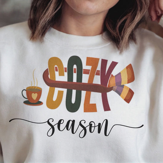 Relaxed woman in white Cozy Season sweatshirt smiles, camera pans to fall leaves, then fades back to show her sipping coffee