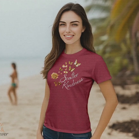 Woman wears Heather Raspberry colored Scatter Kindness T-Shirt while walking outdoors on a tropical beach and enjoying the wind and waves