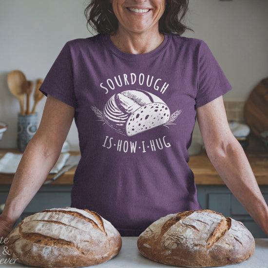 A woman makes a heart sign standing behind sourdough loaves and wearing a Heather Team Purple colored Sourdough Hug T-Shirt