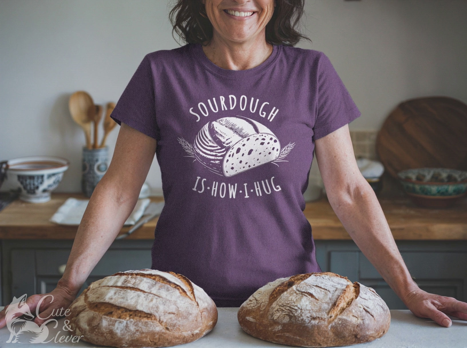 A woman makes a heart sign standing behind sourdough loaves and wearing a Heather Team Purple colored Sourdough Hug T-Shirt