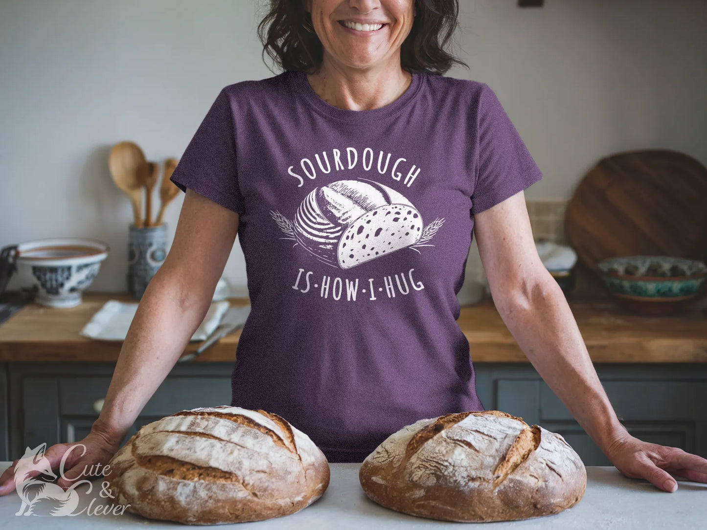 A woman stands in her kitchen behind sourdough loaves wearing a Heather Team Purple colored Sourdough Hug T-Shirt